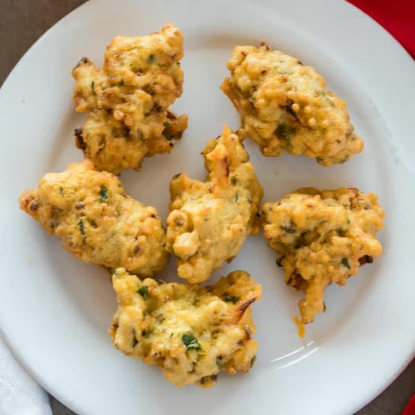 Six golden-brown pakoras, a popular Indian snack, are arranged on a white plate.